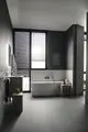 Badewanne mit Wandarmatur und Dusche, Waschbecken mit Spiegel über einer grauen Wand, helle Fliesen und Holzregal; Ideal Standard Produkte sichtbar.